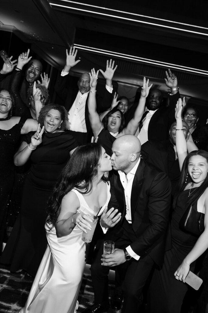 Black and white photo of a bride and groom kissing as they're surrounded by their guests during the reception. 