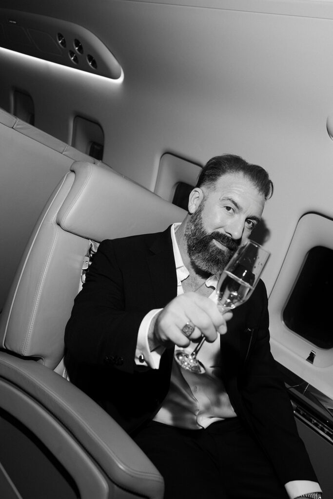 Flash portrait of a groom holding up a glass of champagne as he sits n a private jet for his luxury elopement. 