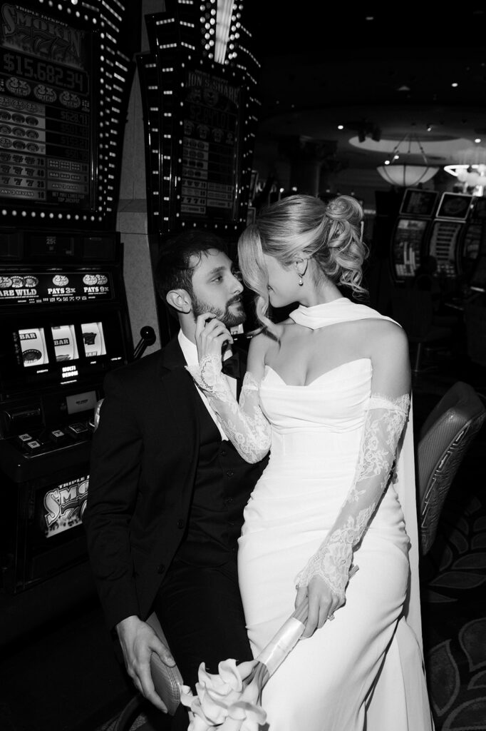 Black and white photo of a bride and groom sitting at the slot machines inside the Caesars Palace casino in Las Vegas. 