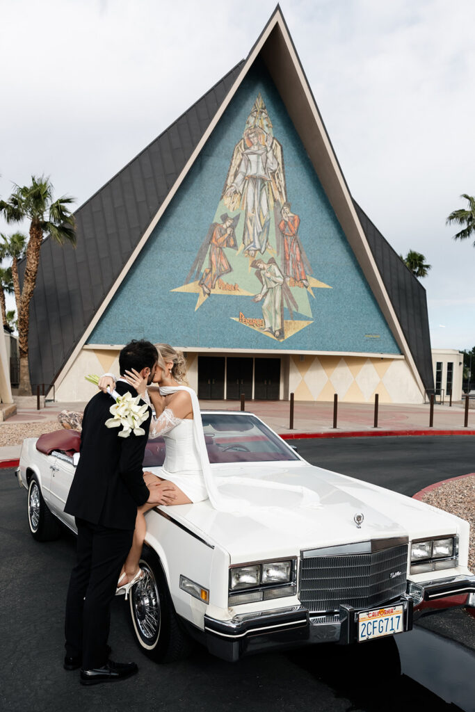 Bride and groom kissing while sitting on a classic convertible outside of Guardian Angel Cathedral in Las Vegas.