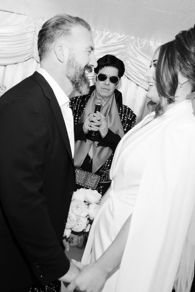 Black and white photo of a bride and groom smiling as they hold hands at the altar with their Elvis officiant. 