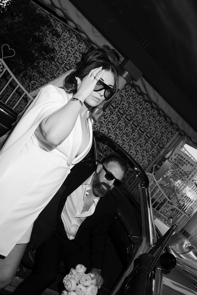 Flash black and white portraits of a bride and groom wearing sunglasses and posing with the pink Cadillac at The Little White Wedding Chapel. 