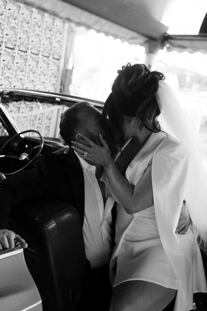 Black and white photo of a bride sitting on the grooms lap as they kiss in a classic convertible. 
