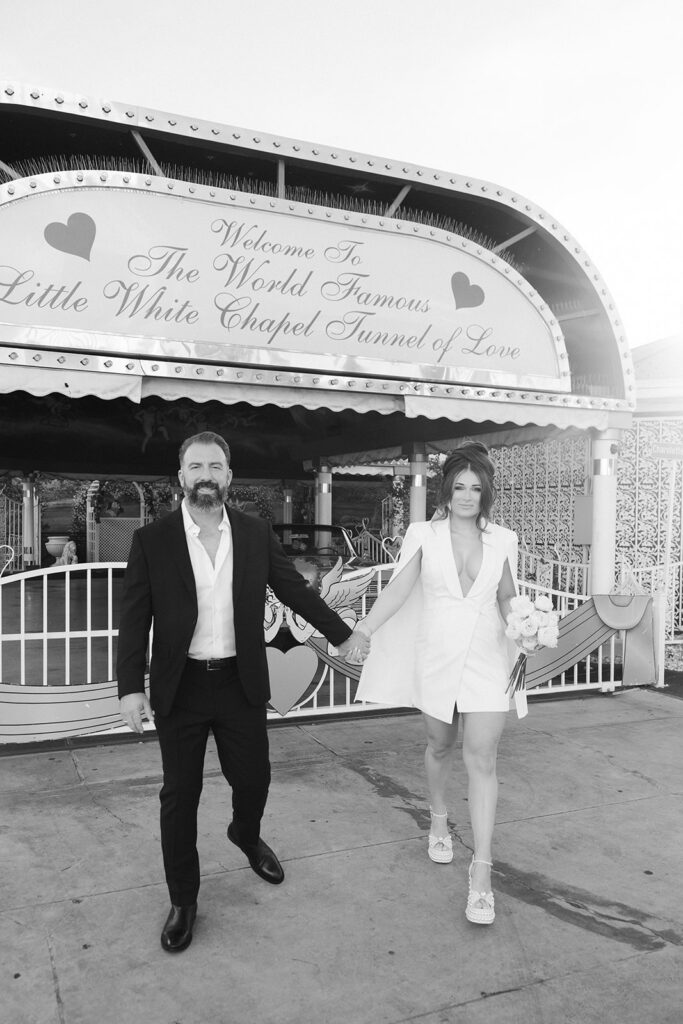 Black and white photo of a bride and groom holding hands outside the Tunnel of Love entrance at The Little White Wedding Chapel in Las Vegas.