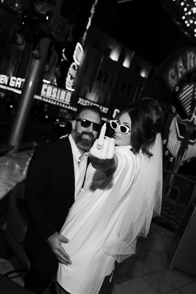 Bride playfully holding up her middle finger while groom sits beside her on Fremont Street in Las Vegas.