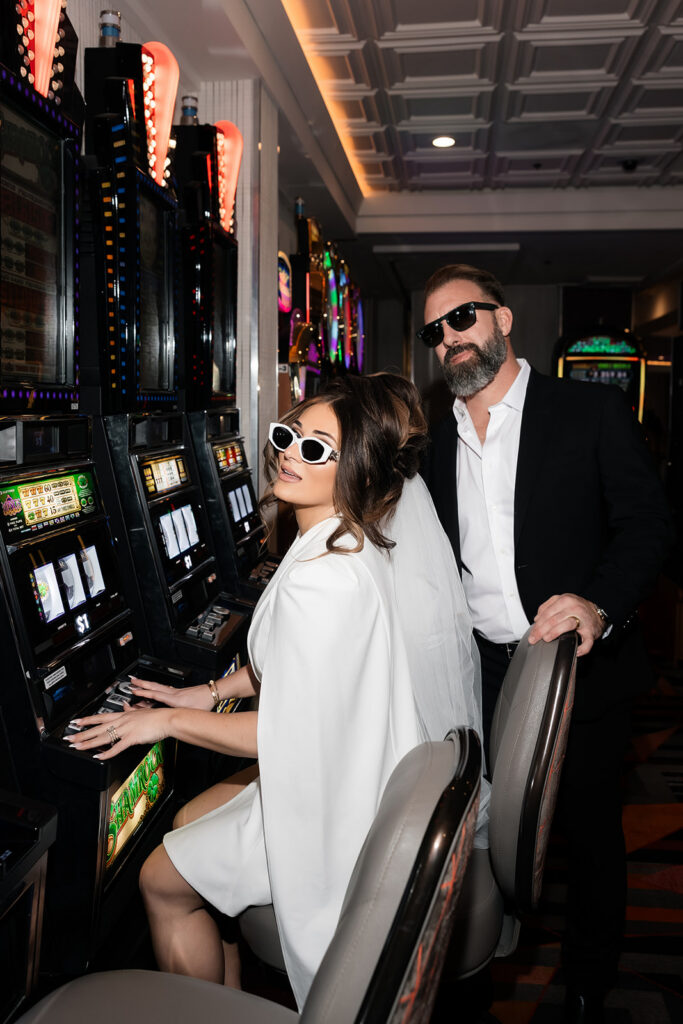 Bride playing a slot machine while groom stands behind her in a Las Vegas casino during their luxury elopement.