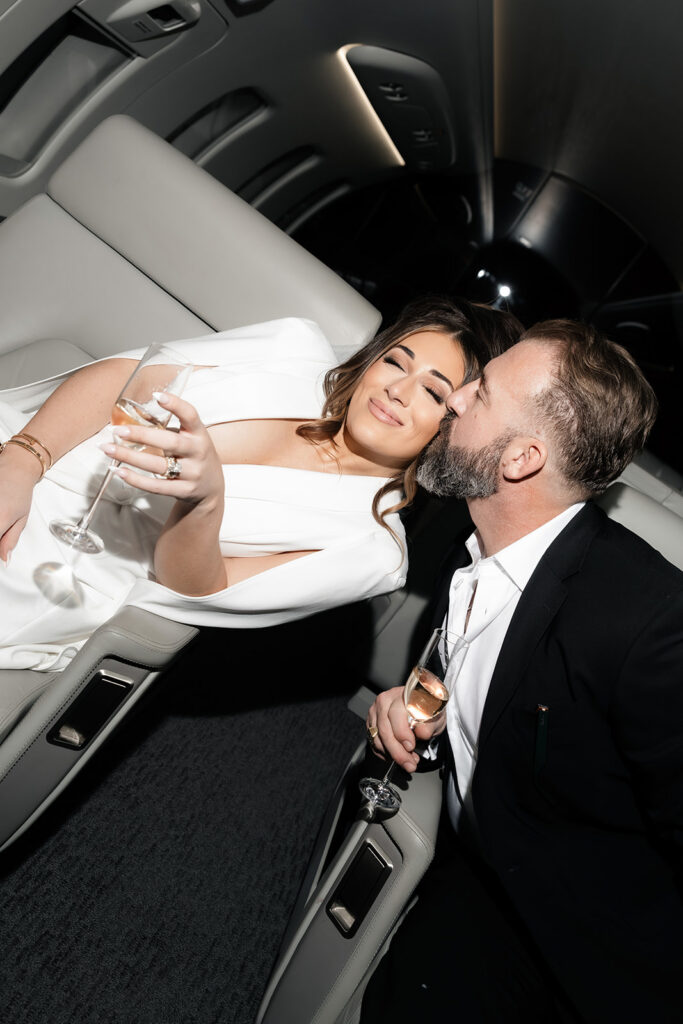 Groom kissing the bride on the cheek while they relax with champagne inside a private jet during their Las Vegas luxury elopement.