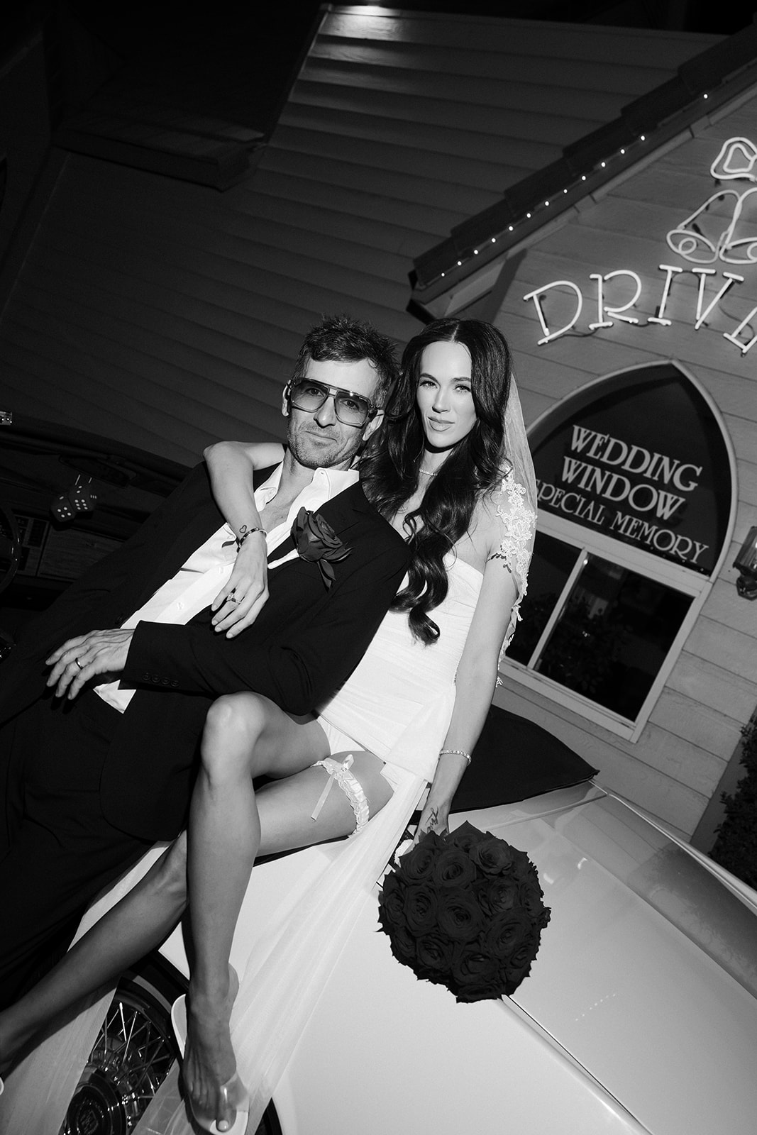 Black and white flash photo of a bride and groom posing with a red convertible outside of A Special Memory Wedding Chapel.