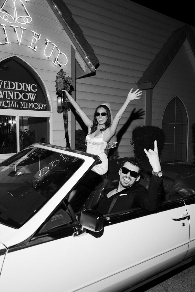 Black and white photo of a couple posing in The Risky Ride outside of the drive-thru wedding window.