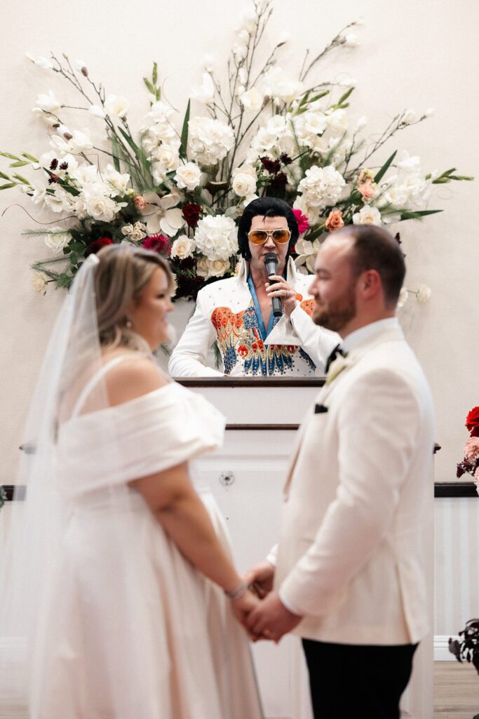 Bride and groom holding hands at the altar while the Elvis impersonator gives a speech during the ceremony.