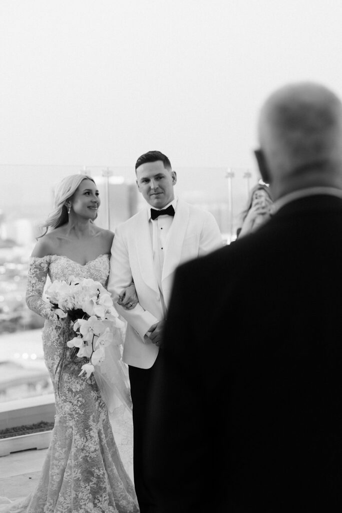 Black and white photo of the bride and groom smiling as the grooms parents give a speech.
