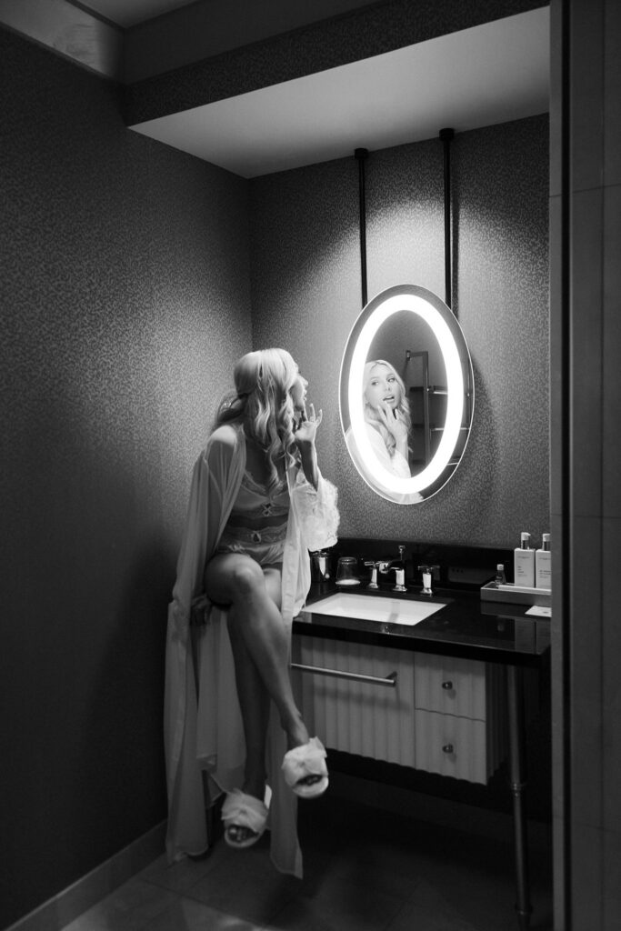 Bride getting ready in a robe while looking into a lighted bathroom mirror at The Cosmopolitan Las Vegas.