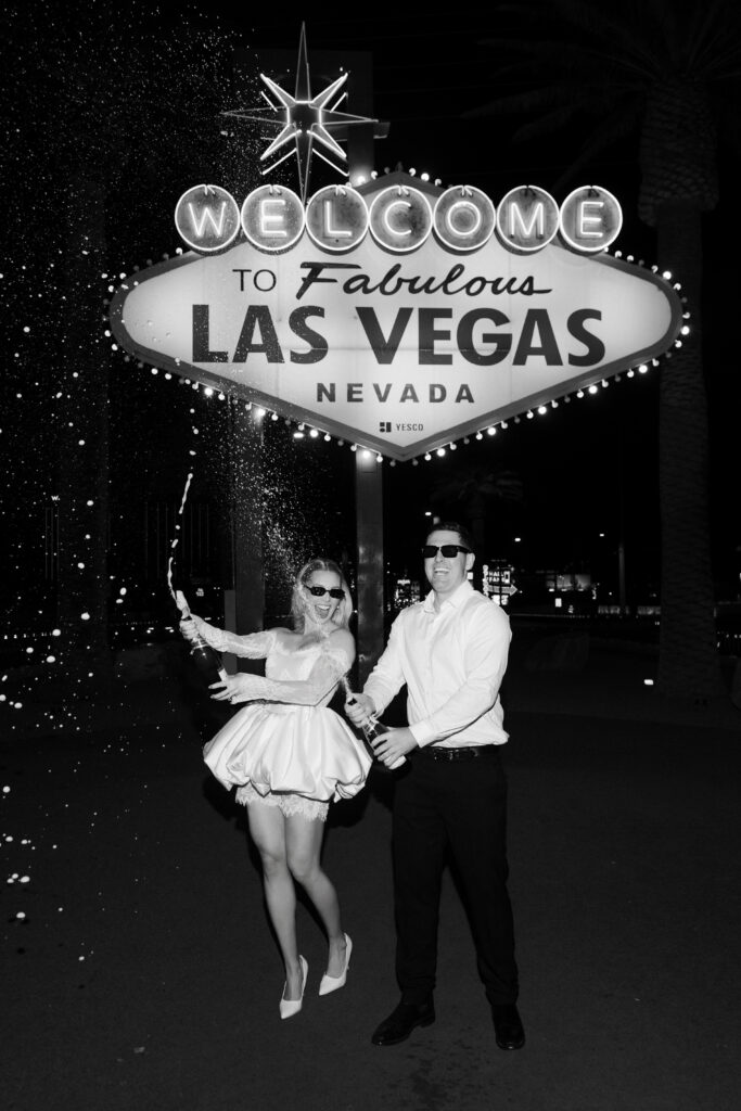 Black and white photo of a couple popping champagne in front of the Welcome to Fabulous Las Vegas sign.