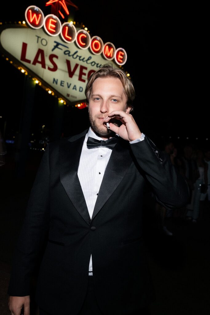 Flash photo of a groom smoking a cigar in front of the Welcome to Fabulous Las Vegas sign at night,
